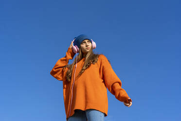 Porträt eines Teenagers vor blauem Himmel, der mit Kopfhörern Musik hört - ERRF02708