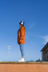 Teenage girl standing on wall against sky listening music with headphones - ERRF02709