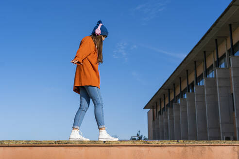 Teenage girl balancing on a wall against while listening music with headphones - ERRF02711