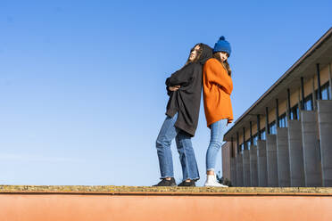 Zwei Teenager-Mädchen stehen Rücken an Rücken an einer Wand vor blauem Himmel - ERRF02716