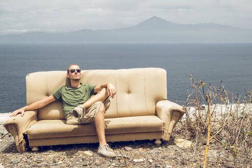 Young man relaxing on broken couch, enjoying the sun, La Gomera, Spain - MAMF01219