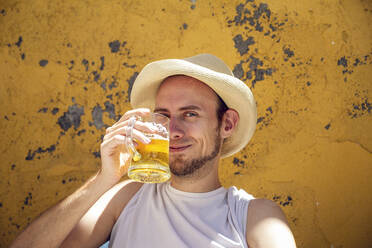 Young man sitting in front of flaking yellow wall, drinking beer, toasting - MAMF01221