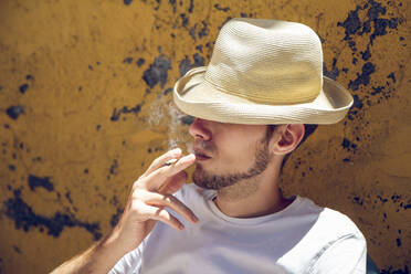 Junger Mann sitzt vor einer abblätternden gelben Wand und raucht eine Zigarette - MAMF01222