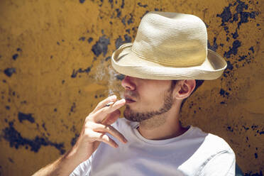 Young man sitting in front of flaking yellow wall, smoking a cigarette - MAMF01222