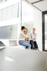 Businesswomen sitting on windowsill in office building, taking a break, talking - PESF01842