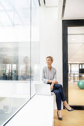 Blond businesswoman with laptop, sitting on windowsill in office building - PESF01845