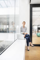 Blond businesswoman with laptop, sitting on windowsill in office building - PESF01846