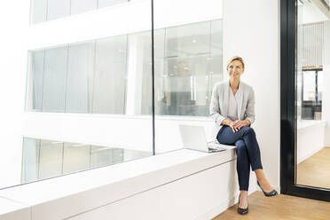 Blonde Geschäftsfrau mit Laptop, die auf der Fensterbank eines Bürogebäudes sitzt - PESF01847