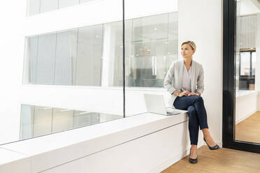 Blond businesswoman with laptop, sitting on windowsill in office building - PESF01848