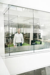 Mature businessman looking out of office building, holding laptop - PESF01850