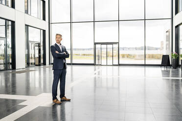 Successful businessman standing in entrance hall of office building, with arms crossed - PESF01860
