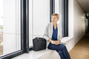 Blonde Geschäftsfrau mit Laptop, die auf der Fensterbank eines Bürogebäudes sitzt - PESF01863