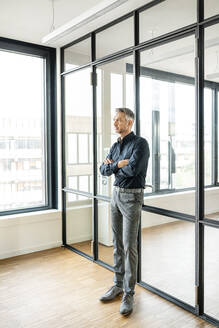 Geschäftsmann lehnt mit verschränkten Armen an einer Glaswand in seinem Büro - PESF01881