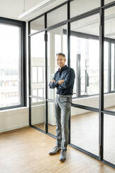 Businessman leaning on glass wall in his office, with arms crossed - PESF01882