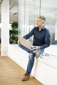 lässiger Geschäftsmann, der auf der Fensterbank eines Bürogebäudes sitzt und einen Laptop benutzt - PESF01890
