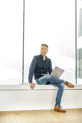 casual businessman sitting on windowsill in office building, using laptp - PESF01893