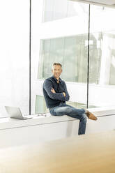 casual businessman sitting on windowsill in office building, with laptop next to him - PESF01896