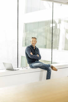 lässiger Geschäftsmann auf der Fensterbank eines Bürogebäudes sitzend, mit Laptop neben sich - PESF01896
