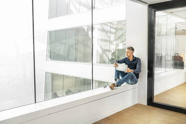 Casual businessman sitting on windowsill in office building, using laptp - PESF01897