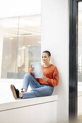 Entspannte Geschäftsfrau, die auf der Fensterbank eines Bürogebäudes sitzt und ein digitales Tablet benutzt - PESF01909