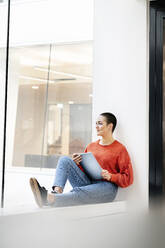 Relaxed businesswoman sitting on windowsill in office building, using digital tablet - PESF01910