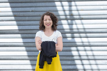 Portrait of smiling brunette woman at roller shutter - HBIF00052