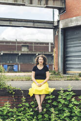 Frau sitzt auf einer mit Pflanzen bewachsenen Mauer in einem alten Industriegebiet - HBIF00055