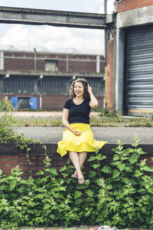 Glückliche Frau sitzt auf einer mit Pflanzen bewachsenen Mauer in einem alten Industriegebiet - HBIF00056