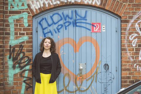 Porträt einer brünetten Frau, die vor einer Backsteinmauer mit einem alten Tor und Graffiti steht, lizenzfreies Stockfoto