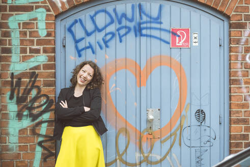 Porträt einer glücklichen brünetten Frau, die vor einer Backsteinmauer mit einem alten Tor und Graffiti steht - HBIF00061