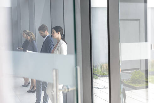 Geschäftsleute, die in einem Bürogebäude warten und sich an eine Glasscheibe lehnen - BMOF00250