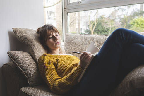 Porträt einer auf der Couch liegenden Frau, die in einem Skizzenbuch zeichnet - WPEF02673