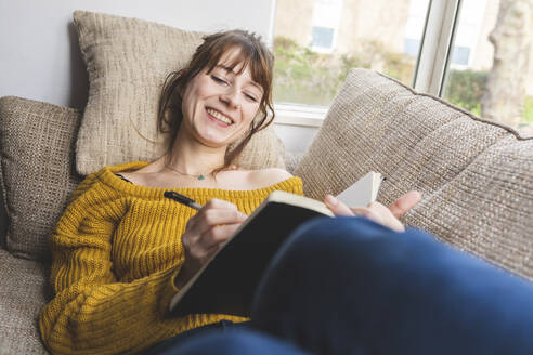 Porträt einer glücklichen Frau, die auf der Couch liegt und in ein Skizzenbuch zeichnet - WPEF02674