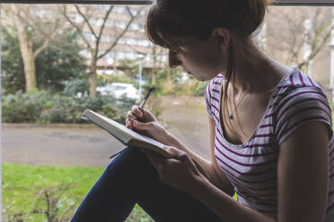 Woman sitting in front of windowpane drawing in sketchbook - WPEF02675