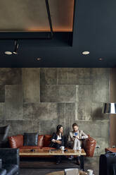 Businessman and businesswoman sitting on couch in hotel lobby sharing smartphone - ZEDF03019