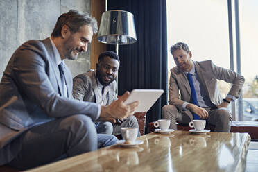 Three businessmen sitting in hotel lobby with tablet - ZEDF03037