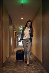 Businesswoman walking in hotel corridor with rolling suitcase - ZEDF03105