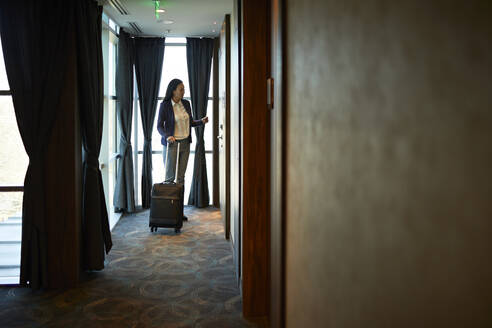 Businesswoman in hotel corridor with rolling suitcase - ZEDF03106