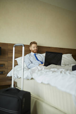 Geschäftsmann auf dem Bett liegend in einem Hotelzimmer mit Laptop, lizenzfreies Stockfoto