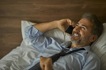 Smiling businessman lying on the bed in hotel room talking on the phone - ZEDF03162