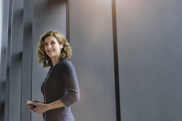 Portrait of blond businesswoman with earphones and smartphone in office - BMOF00271