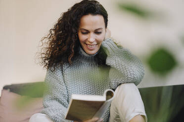 Woman sitting on couch, reading book, smiling - JOSEF00004