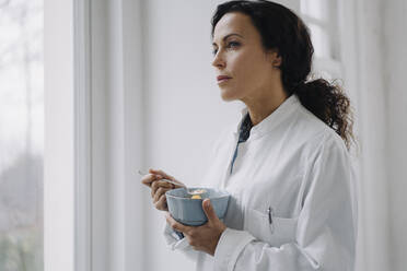 Female doctor eating a snack, standing at window - JOSEF00021