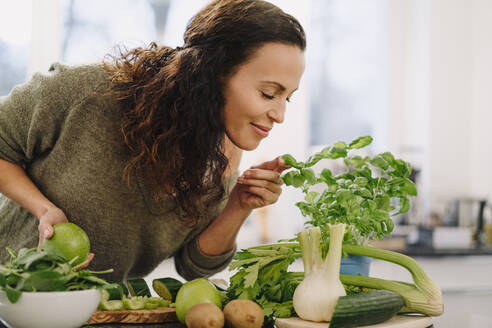 Woman standing in kitchen, smelling ingredients for healthy smoothie - JOSEF00026