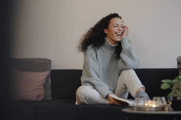 Woman sitting on couch laughing, reading a book - JOSEF00050