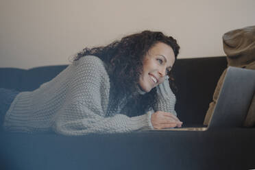 Woman lying on couch, using laptop, smiling - JOSEF00061