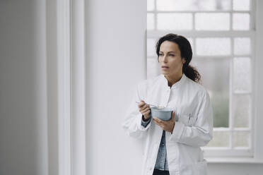 Female doctor eating a snack, standing at window - JOSEF00090