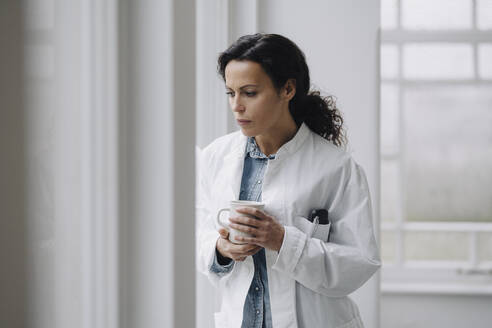 Female doctor standing at the window, looking worried, holding cup of coffee - JOSEF00096