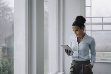 Erfolgreiche Geschäftsfrau, stehend am Fenster, mit digitalem Tablet, lesend - JOSEF00098