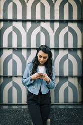 Portrait of young woman with long black hair standing in front of patterned wall using smartphone - DCRF00025